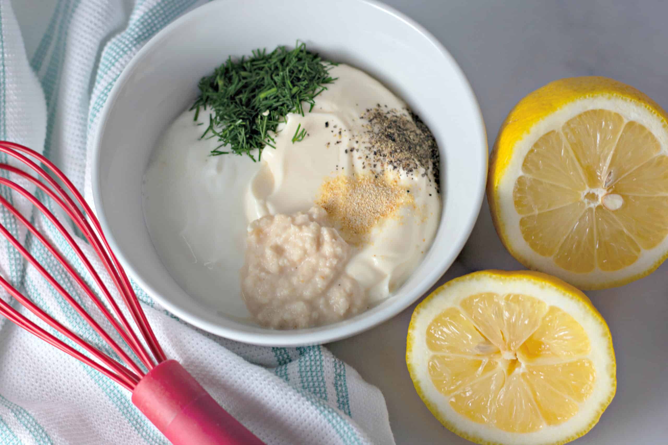 Creamy Dill Sauce ingredients in a white bowl with a red whisk and sliced lemons